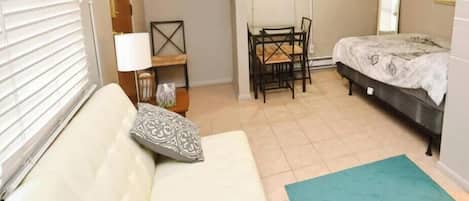 Une pièce comprenant un lit, un canapé, une table avec des chaises, un téléviseur fixé au mur et une fenêtre avec des stores.