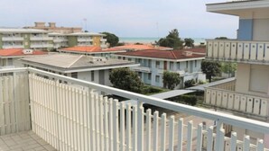 Ein Balkon mit weißer Brüstung, der auf ein Wohngebiet mit Gebäuden und einen Strand im Hintergrund blickt.