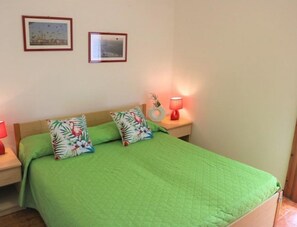 Ein Schlafzimmer mit grüner Bettdecke, zwei Kissen mit tropischen Motiven, einer Nachttischlampe und zwei gerahmten Bildern an der Wand.