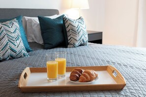 A wooden tray with two glasses of orange juice and two croissants on a bed with a grey quilted cover. - Hampden Apartments - The Charles (Windsor)