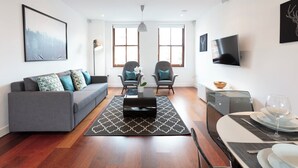 A modern living room with a grey sofa, two armchairs, a coffee table, and a dining area with a glass table and chairs. - Hampden Apartments - The Charles (Windsor)