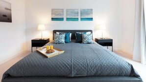 A modern bedroom with a large bed, two bedside tables with lamps, a tray with a croissant and juice, and three paintings on the wall. - Hampden Apartments - The Charles (Windsor)