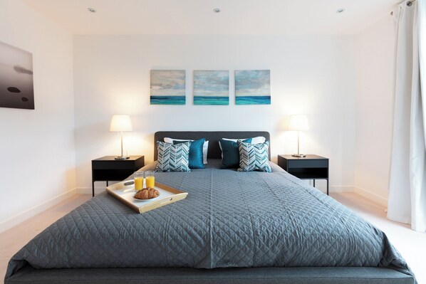 A modern bedroom with a large bed, two bedside tables with lamps, a tray with a croissant and juice, and three paintings on the wall. - Hampden Apartments - The Charles (Windsor)