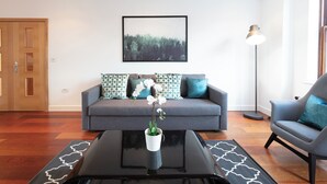 A modern living room with a grey sofa, patterned cushions, a black coffee table, and a framed landscape painting on the wall. - Hampden Apartments - The Charles (Windsor)