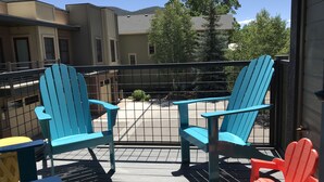 A balcony with two blue Adirondack chairs and one red Adirondack chair, a wooden deck, and a view of buildings and trees.