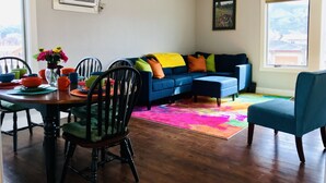 A living room with a blue sofa, colorful cushions, a dining table with chairs, a chandelier, and a ceiling fan.