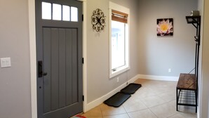 A hallway with a gray door, a window with blinds, a decorative wall piece, a mat, and a small table with a metal frame.