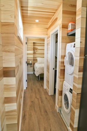 A narrow hallway with wooden walls and floors, leading to a bedroom with a bed and a washing machine.