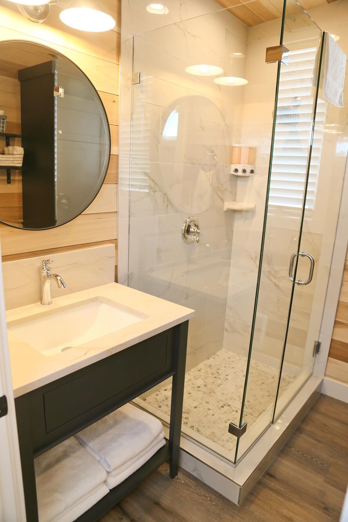 Een moderne badkamer met een douchecabine van glas, een ronde spiegel en een witte wastafel.