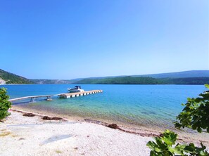 Un lago tranquilo con un muelle de madera que se adentra en el agua, rodeado de una playa de guijarros y vegetación.