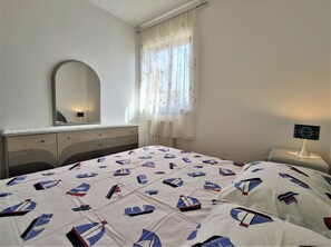 Un dormitorio con cama, cómoda, espejo, ventana con cortinas y lámpara de noche.