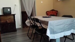 Ein Essbereich mit einem Tisch, der mit einer tischdurchmusterter Tischdecke bedeckt ist, umgeben von vier Stühlen. Ein Fernseher steht auf einer Holzkommode links.