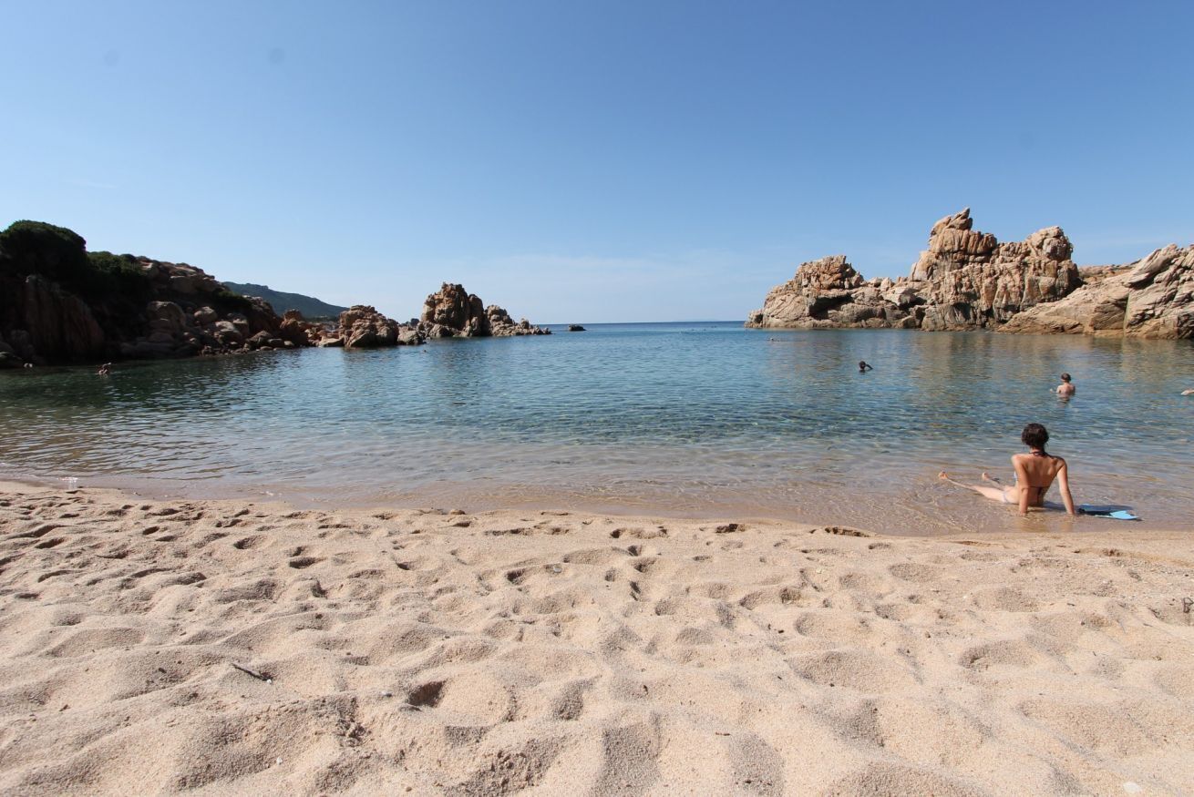 A sandy beach with rocky formations in the background and clear blue water.