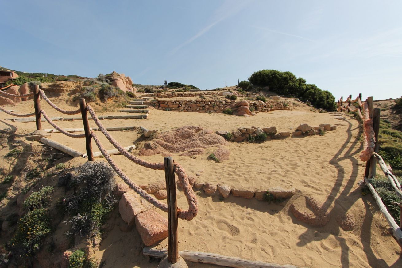 Un site archéologique sablonneux, avec des structures en pierre et une clôture en bois.