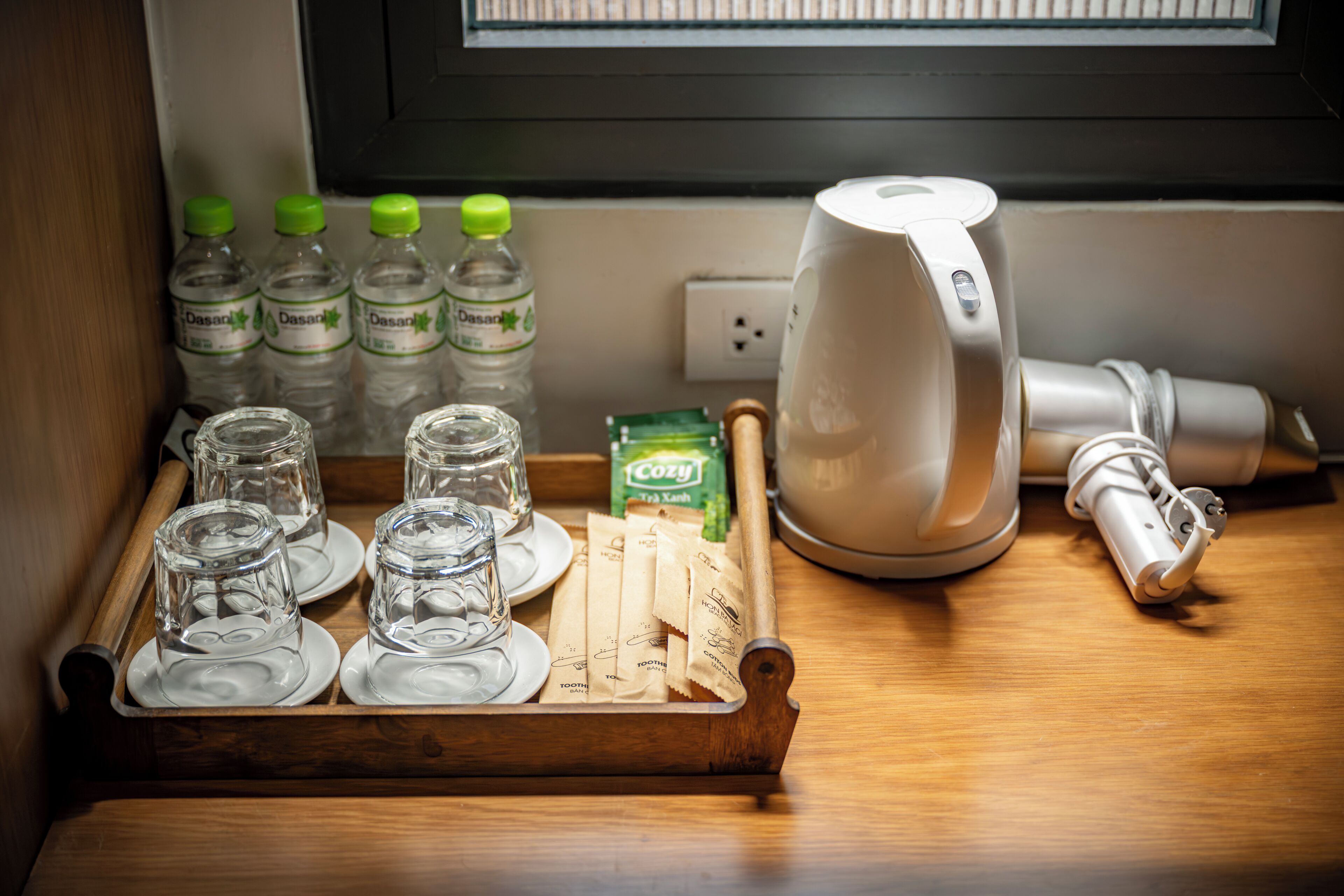A tray with glasses, Dasani water bottles, and tea packets on a wooden surface near a window.