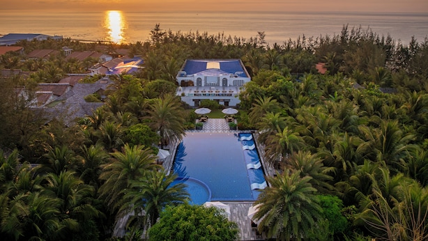 A resort with a swimming pool, surrounded by palm trees, and a view of the ocean at sunset. - Honba Lagi Beach Resort (La Gi)
