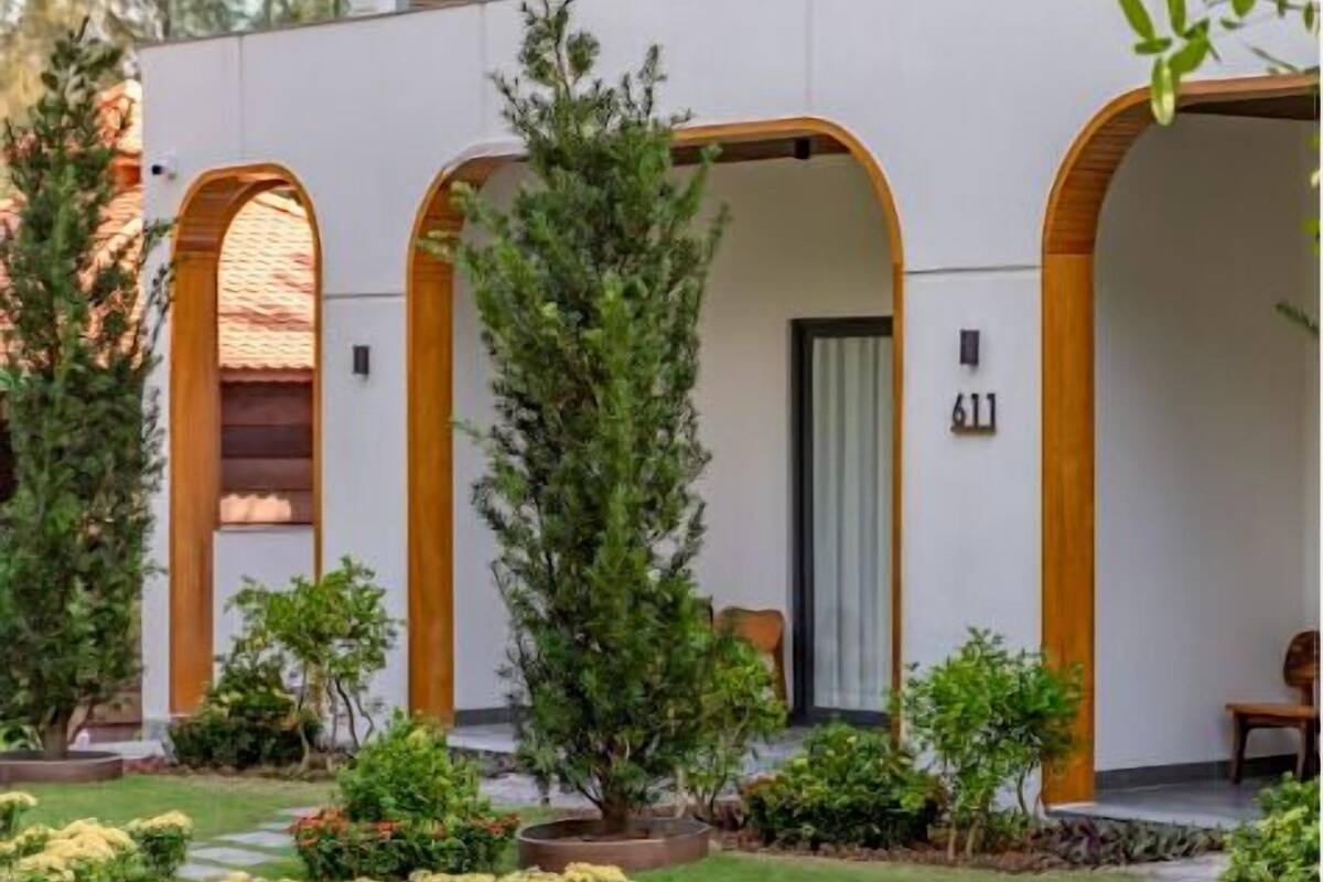 Un edificio moderno con fachada blanca, arcos de madera y un jardín con diversas plantas.