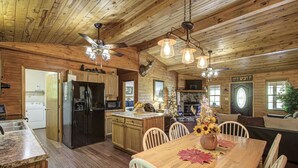 Interior rústico de una cabaña con paredes de madera, un comedor con mesa y sillas de madera, una cocina con refrigerador y estufa, y un ventilador de techo.