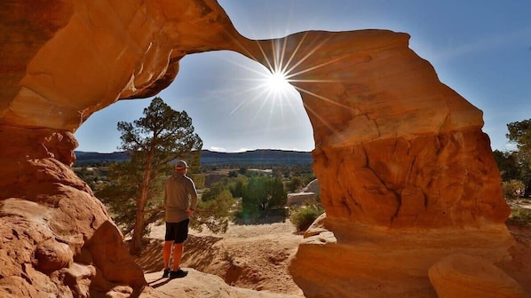 A person standing under a natural rock arch with the sun shining through, surrounded by a landscape with trees and hills.