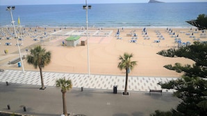 Ein Strand mit Reihen blauer Sonnenschirme und Liegen, ein von Palmen gesäumter Weg und freie Sicht auf das Meer.