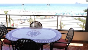 Ein Balkon mit rundem Tisch und Stühlen, der Blick auf einen Strand mit Personen und Booten geht.
