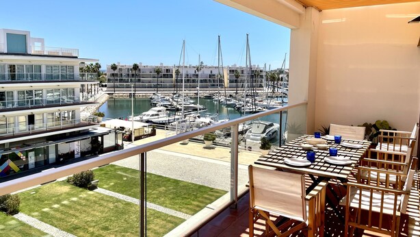 A balcony with a view of a marina, a dining table set with blue glasses, and a potted plant.