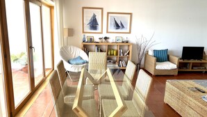A spacious living area with a glass-top dining table surrounded by wicker chairs, a sofa, and a television.