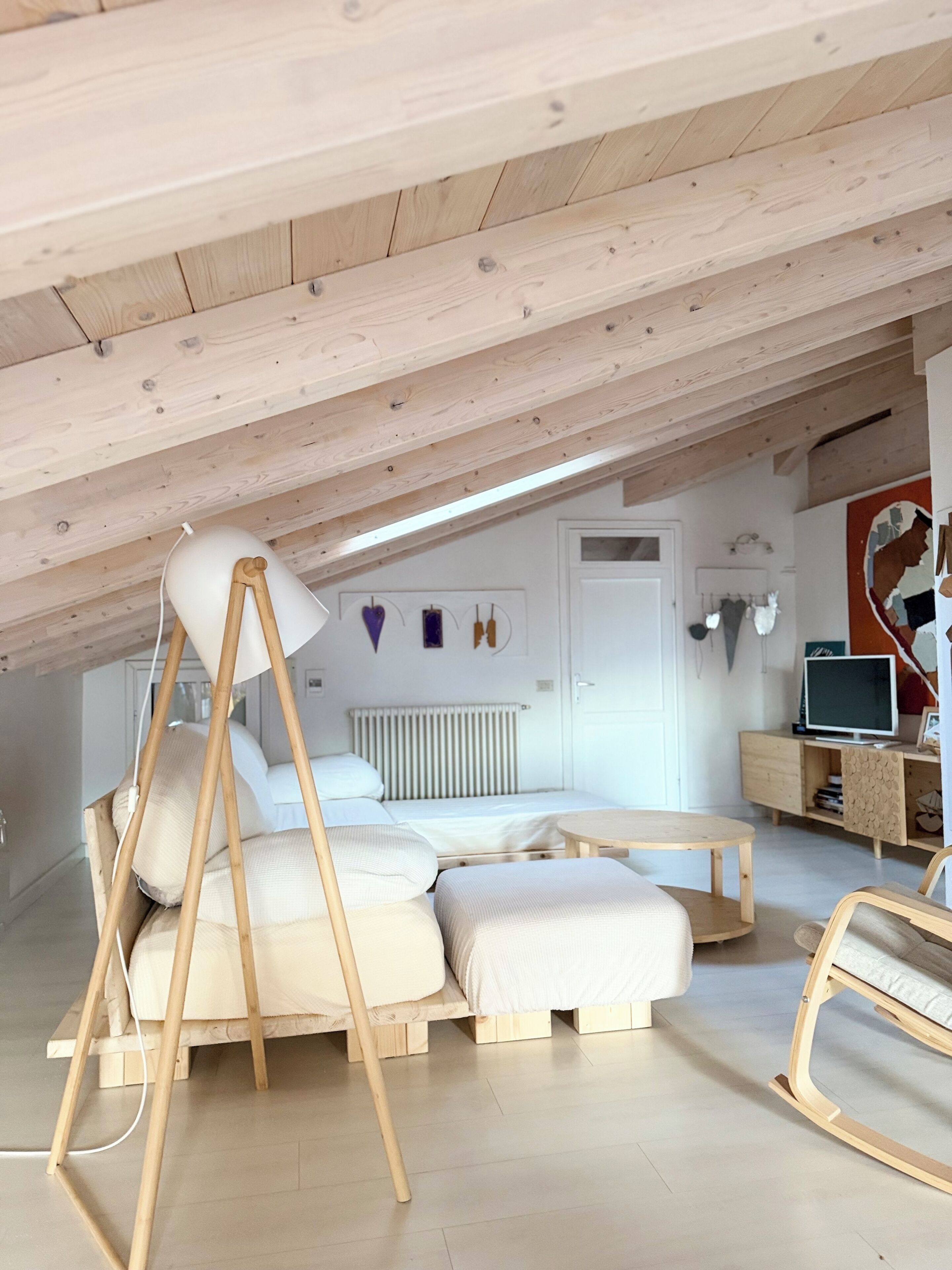 Une chambre à coucher aménagée dans un grenier, avec un plafond en pente, des poutres en bois, un lit avec un cadre en bois, une table de chevet, une chaise et un téléviseur posé sur un meuble en bois.