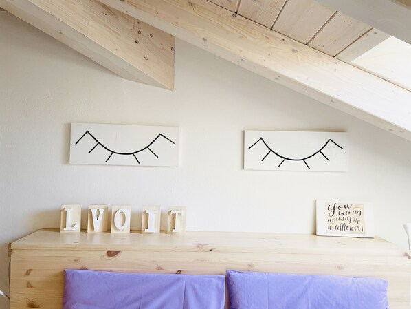 Ein Zimmer mit schräger Decke, Holzbalken, einem Bett mit violetter Bettdecke sowie Wanddekorationen, darunter zwei abstrakte Kunstwerke und ein gerahmter Spruch.