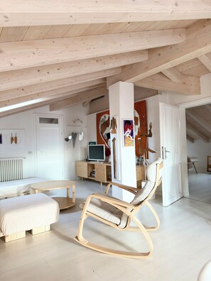 Ein großzügiges Loft mit hohen Decken, freiliegenden Holzbalken, einem Schaukelstuhl, einem Bett, einem Schreibtisch mit Computer und einem großen Fenster.