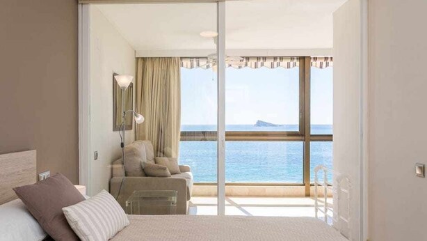 Hotelkamer met een bed, een slaapbank, een stoel, een lamp en uitzicht op zee door grote ramen.