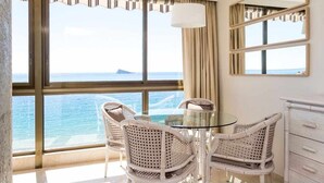 Een kamer met een glazen tafel en rieten stoelen, een groot raam met uitzicht op zee, en een witte linnenkast met spiegel.