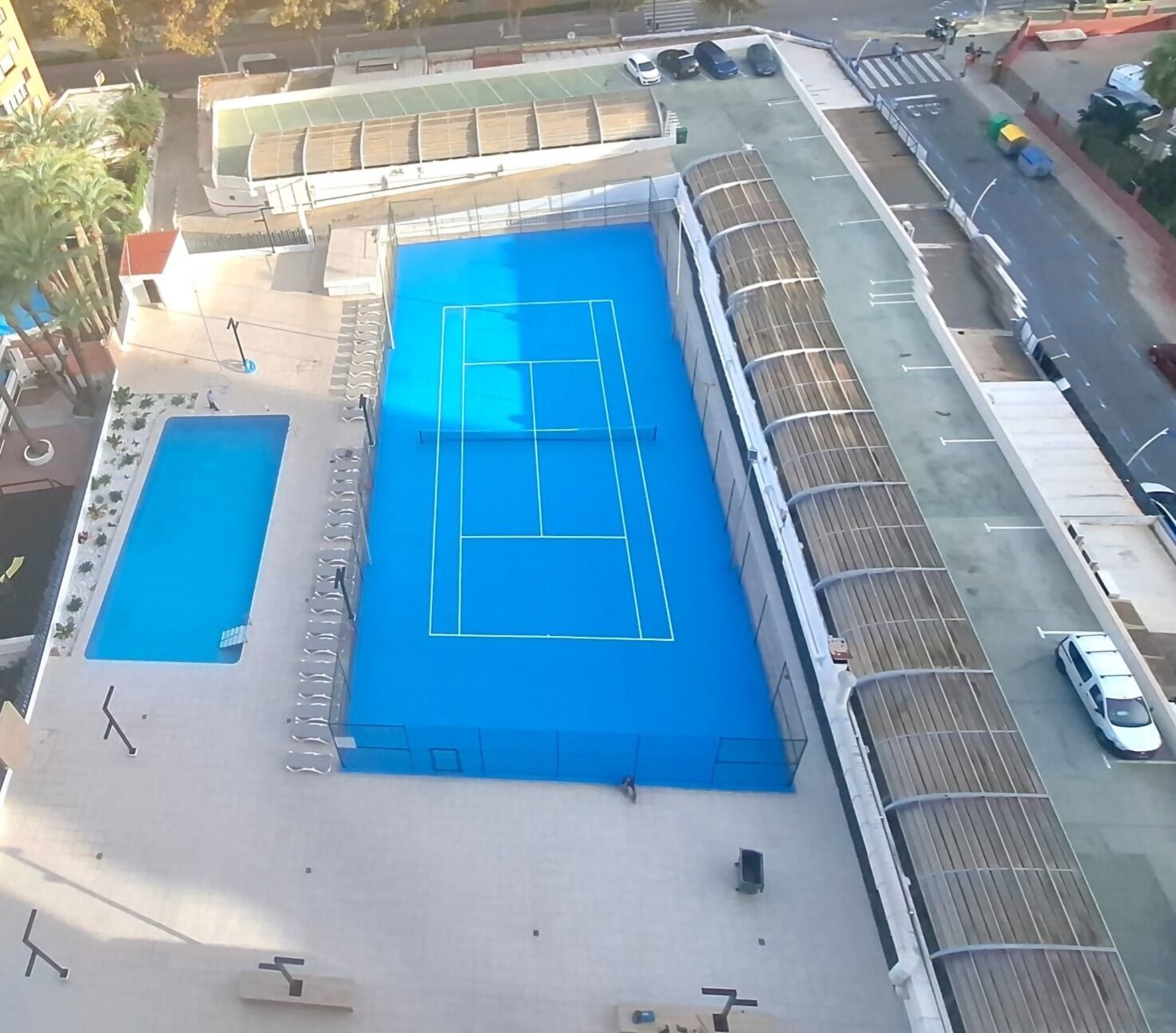 Zwembad op het dak met een tennisbaan, omgeven door een hoge afrastering en een parkeerplaats.