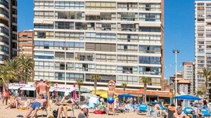 Een hoog hotel met meerdere balkons, direct aan het strand, met een zandstrand vol mensen en parasols.