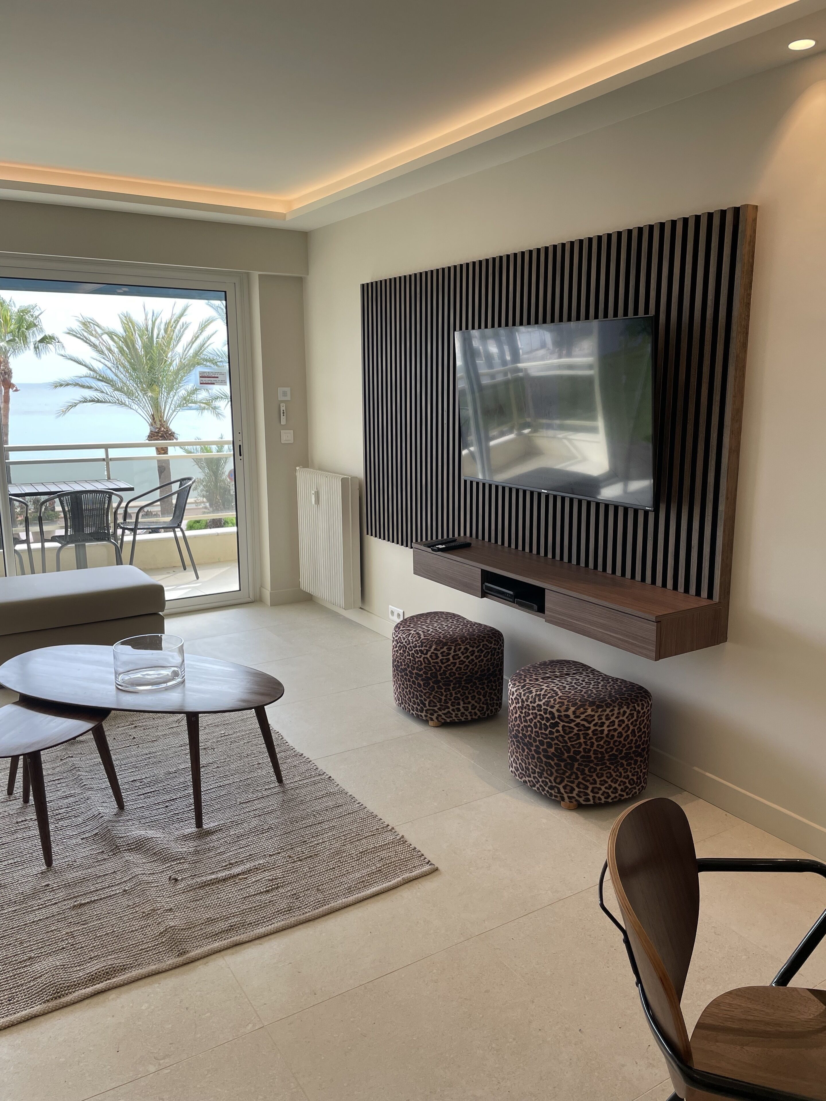 A modern living room with a large TV mounted on a wall, a round coffee table, two leopard print ottomans, and a view of the sea through a sliding glass door.