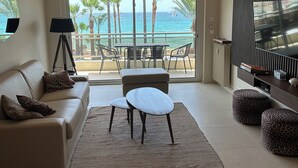 Un salon moderne comprenant un canapé, deux fauteuils, une table basse et un téléviseur. La pièce est dotée d’une grande porte-fenêtre coulissante qui donne sur un balcon offrant une vue sur l’océan et les palmiers.
