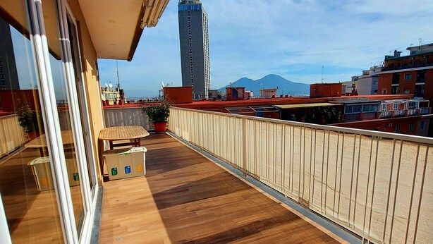 A balcony with wooden flooring, a potted plant, and a view of a tall building and mountains in the distance. - SUITE WITH VESUVIUS VIEW TERRACE IN HISTORIC CENTER (Napoli)