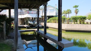 Un muelle de madera se extiende sobre una superficie acuática tranquila, con una zona cubierta y un edificio con balcón al fondo.