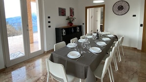 A dining area with a marble floor, a long table set for six, white chairs, a black cabinet, and a wall clock. - The terrace - vacation home with lake view (San Zeno di Montagna, Verona)