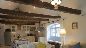A cozy living room with a sofa and a dining table, a kitchen with wooden beams, and a window with curtains.