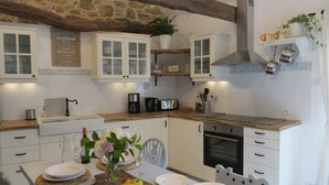 A modern kitchen with white cabinets, a stone wall, and a wooden ceiling. There is a dining table set with plates, a fruit bowl, and a vase with flowers.