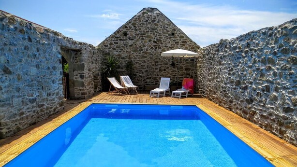 A swimming pool with stone walls and wooden decking, surrounded by lounge chairs and an umbrella.