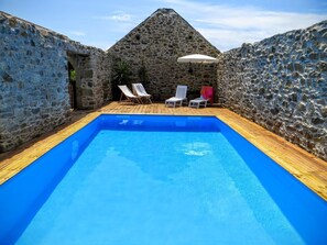 Ein Pool mit Steinwänden und Holzterrasse, umgeben von Liegestühlen und einem Sonnenschirm.