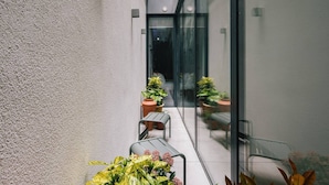 Een smalle buitenpassage met een glazen deur die uitkomt op een terras. Er staan potplanten en tuinmeubilair langs de passage.