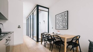 Een moderne keuken met een ingebouwde kookplaat, een eethoek met een houten tafel en zwarte stoelen, en een grote glazen deur die leidt naar een lichte kamer.
