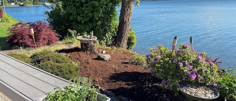 Una terraza con vista a un lago, rodeada por un jardín con diversas plantas y un bebedero para pájaros.