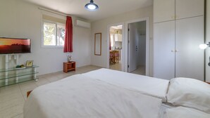 Een slaapkamer met een groot bed, een tv op een standaard, een klein tafeltje met een plant en een raam met gordijnen.