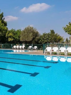 Piscina com água azul cristalina, cercada por espreguiçadeiras brancas e cercas verdes.