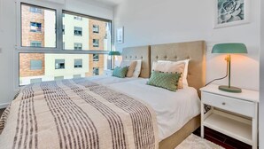 Ein modernes Schlafzimmer mit einem großen Bett, zwei Nachttischen, einer grünen Lampe und Blick auf ein Backsteingebäude durch große Fenster.