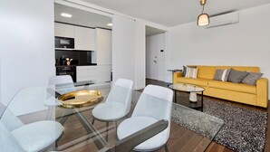 Ein moderner Wohnraum mit einem Glastisch, weißen Stühlen, einem gelben Sofa und einer Küche im Hintergrund.